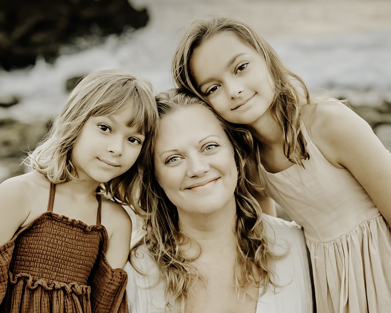 Family Photographer, a mother sits between her two young daughters, they all smile at the beach