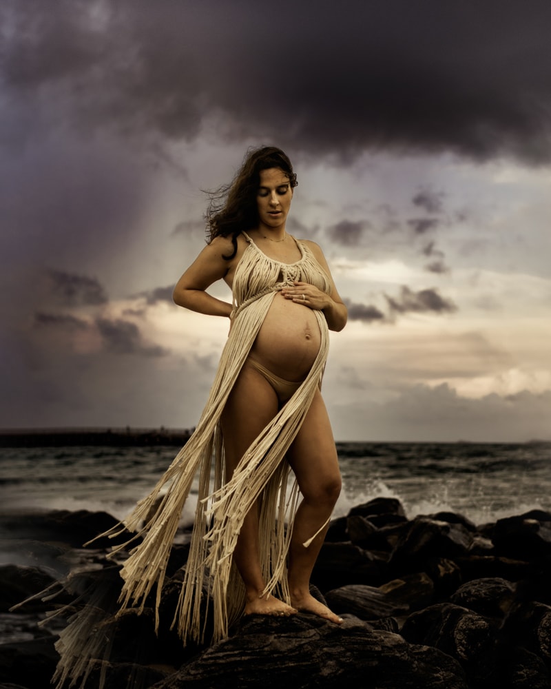 maternity photographer, an expecting mother has her hand on her belly as she stands on rocks at the beach