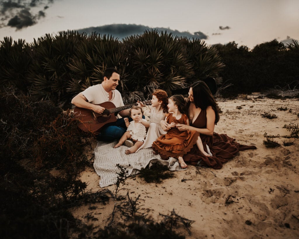 Family Photographer, a father plays the guitar on the beach as his wife and three children sit and listen
